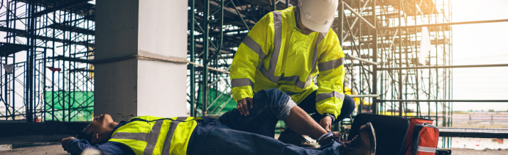 A construction worker lies unconscious on a concrete floor at a construction site while a supervisor or first responder in a high-visibility jacket and hard hat kneels to provide first aid next to an open medical kit.