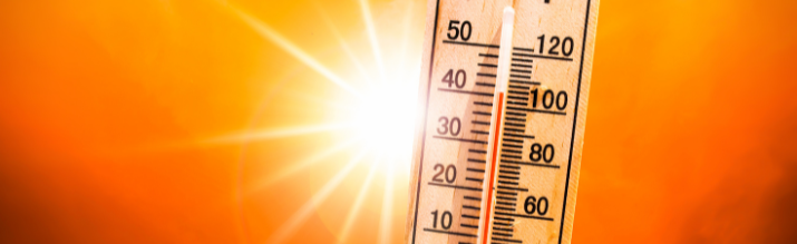 Close-up of a wooden thermometer indicating a high temperature above 100 degrees Fahrenheit against a bright, hazy orange background with an intense sunburst.