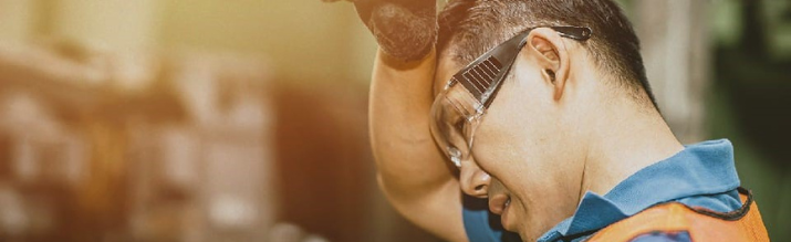 A fatigued industrial worker wearing safety glasses and an orange vest wipes sweat from his forehead in a warm, blurred factory setting.